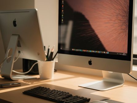 밝은 책상 위에 놓인 게이밍 키보드와 마우스, 화면에 모바일 게임 인터페이스가 표시된 모니터, 흐릿한 창가 배경