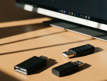 손으로 잡은 스마트폰과 게임 컨트롤러, 노트북 키보드 위에 놓인 이어폰과 USB 케이블, 흐릿한 책상 배경