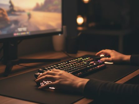 손으로 게이밍 마우스와 키보드를 조작하는 모습, 모니터 화면에 모바일 게임 실행 중, 데스크 조명이 비추는 어두운 방 배경